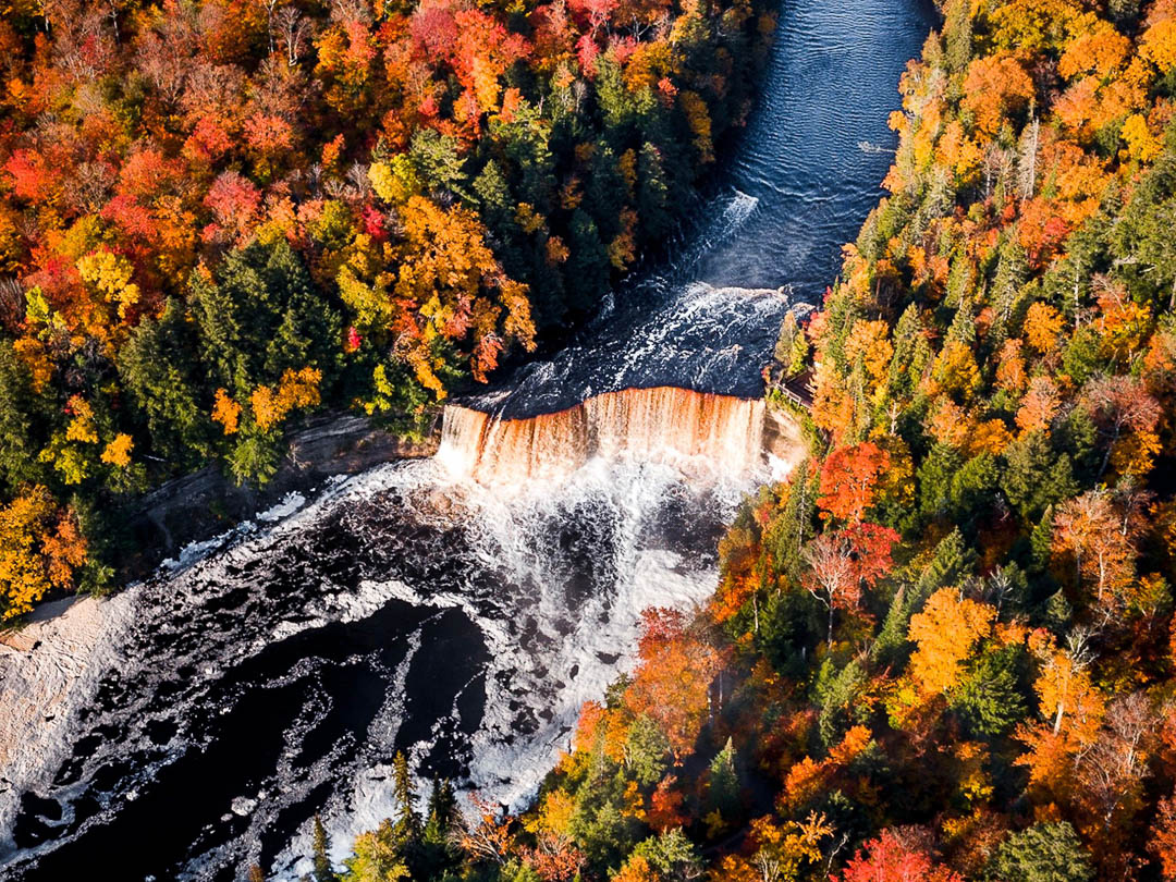 Aerial view of the Upper Tahquamenon Falls pouring over a wide rock ledge ringed by orange and red autumn forest