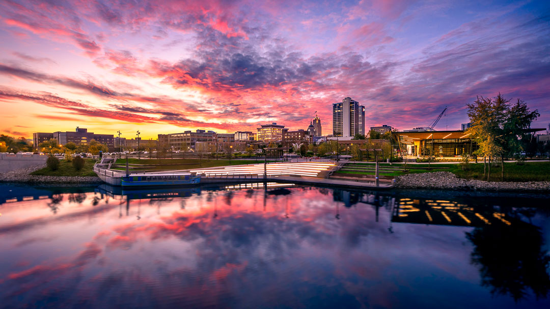 A pink and orange sunset over downtown Fort Wayne reflected in the St. Marys River at Promenade Park