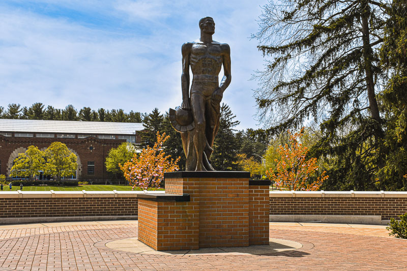 The bronze Spartan statue standing on its brick pedestal under a blue sky on the Michigan State University campus